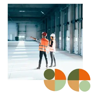 Two facility managers in hard hats and safety vests stand inside a large industrial warehouse, inspecting the space and discussing operations. Represents Air Theory’s support for facility managers and operators with efficient, maintainable, standards-documented mechanical systems.